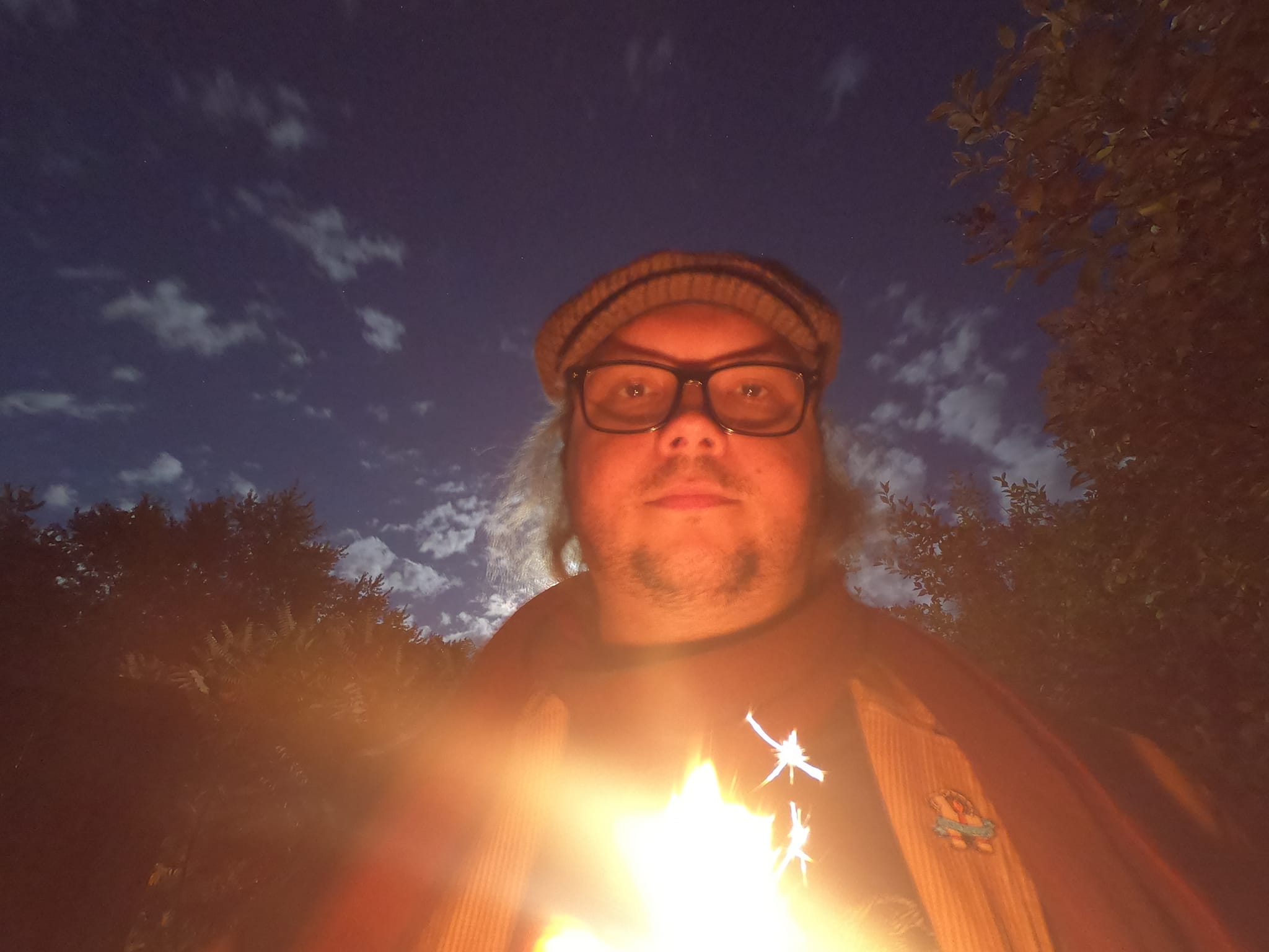 A man with long  hair is holding a sparkler, his features are difficult to make out but is wearing the same clothes as previous pictures. It is dark and the clouds are lit by a bright moon, which is not in frame